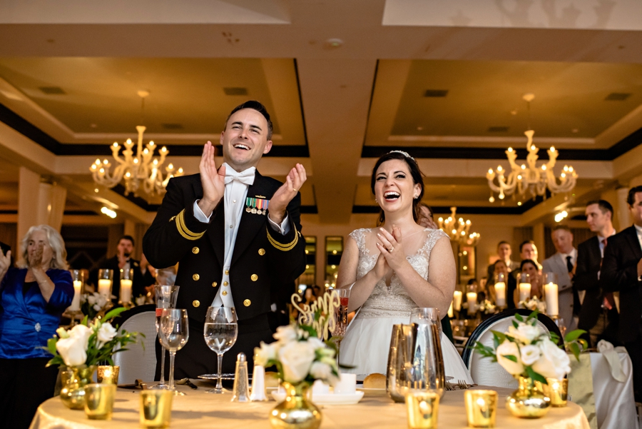 A bride and groom laugh and applaud after a speech.